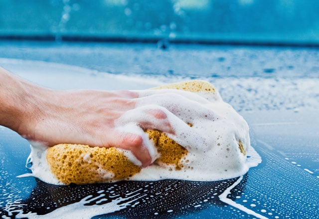 A hand holding a soapy sponge on the bonnet of a blue car